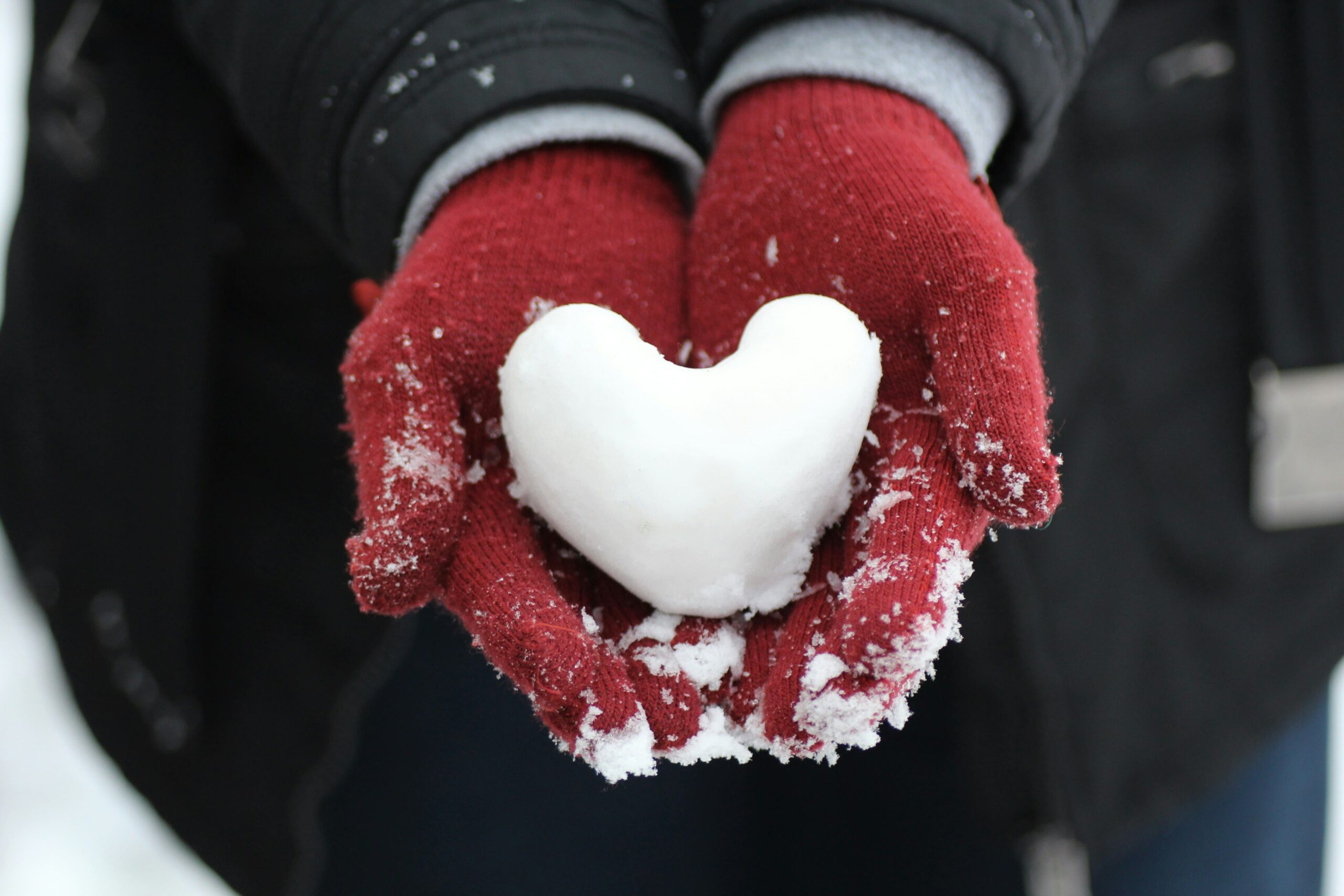 Mãos com luvas segurando uma bola de neve moldada em formato de coração, representando o infarto no inverno.